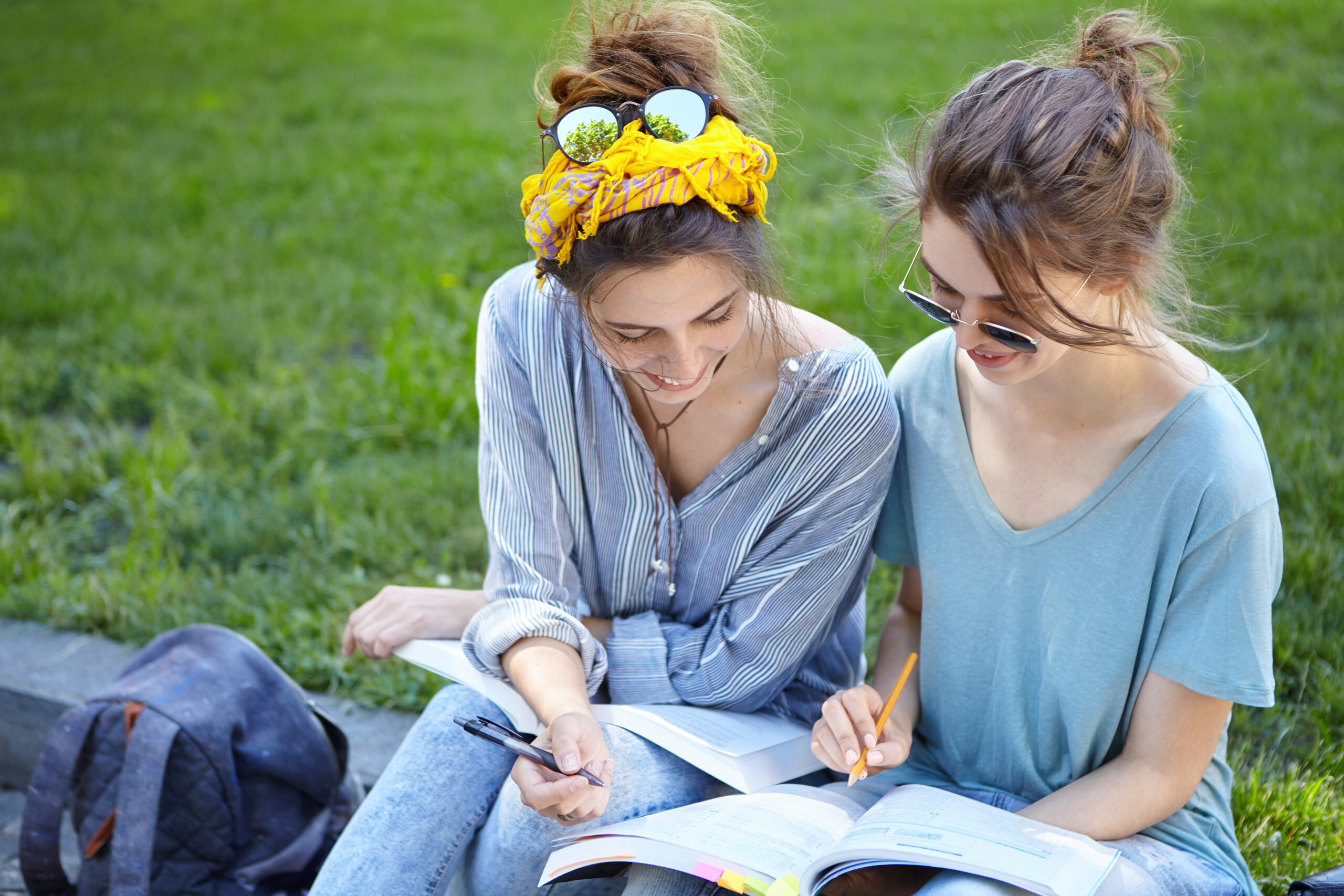Two female students sitting at bench at college campus looking attentively in book writing something down with pencils admiring wonderful nature. Two university friends studying outdoors near lawn