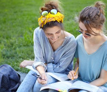 Two female students sitting at bench at college campus looking attentively in book writing something down with pencils admiring wonderful nature. Two university friends studying outdoors near lawn